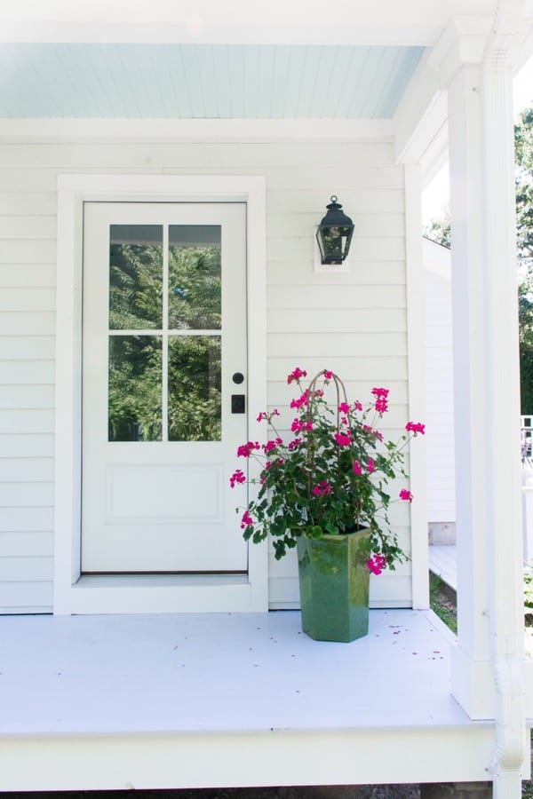 Coastal cottage back porch with blue ceiling and light gray deck with a white door and pink geraniums.