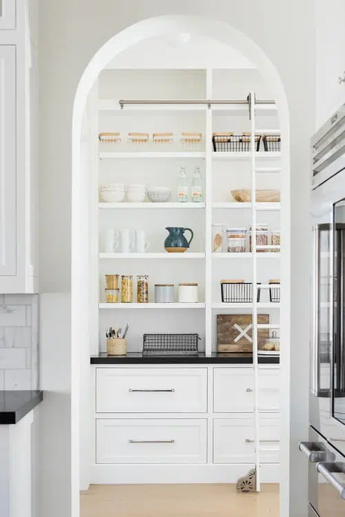 White butler's pantry with black counters, open shelves, ladder and arched doorway