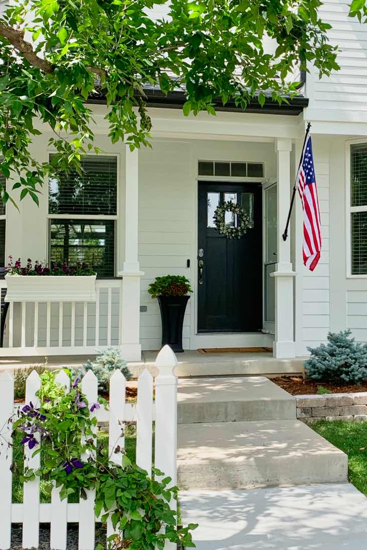 casa blanca con pintura de color blanco de granja, puerta frontal negra, valla blanca y bandera estadounidense