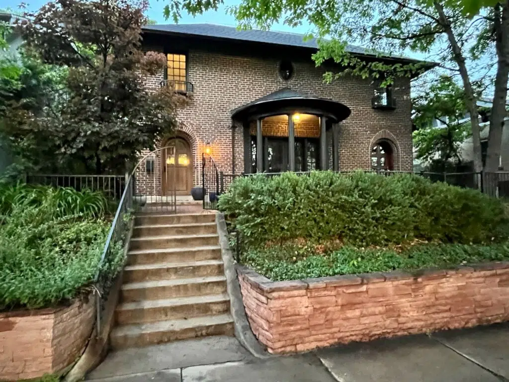 A red brick home with black trim paint is pictured with natural wood front door. 