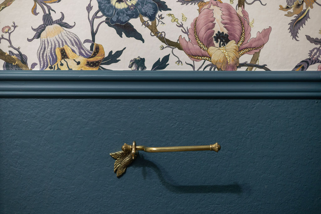 A wall with Bella Blue wainscoting, floral wallpaper and brass toilet paper holder.