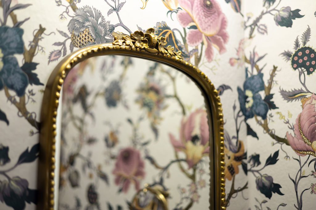 A brass mirror hangs on a wallpaper wall.