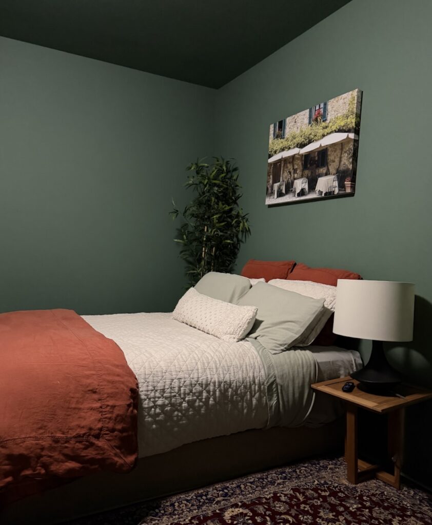 A bedroom drenched with BM Webster Green, with rusty orange bedspread and lights on overhead